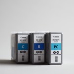 Photograph of three blue Canon photo printer ink cartridges standing upright on a grey background.