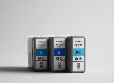 Photograph of three blue Canon photo printer ink cartridges standing upright on a grey background.